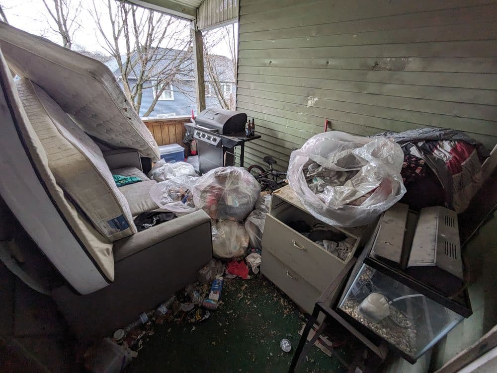 Cluttered porch with trash bags, a mattress, and discarded furniture indicating neglect.